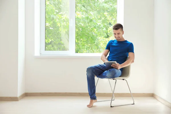 Young man with tablet resting on chair near window at home — Stock Photo, Image