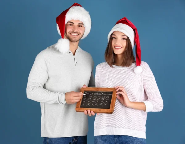 Молодая пара в шляпах Санта-Клауса с доской подсчета дней до Рождества, на цветном фоне — стоковое фото