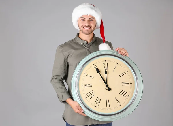 Jeune homme en chapeau de Père Noël avec horloge sur fond gris. Concept de compte à rebours de Noël — Photo