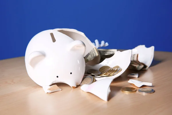 Broken piggy bank with money on table against color background — Stock Photo, Image