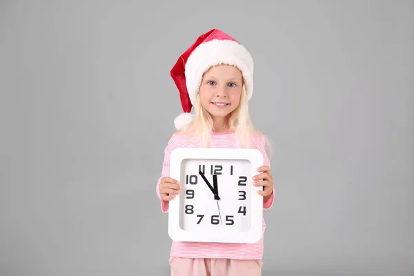 Söt flicka i Tomtemössa med klockan på grå bakgrund. Christmas countdown koncept — Stockfoto
