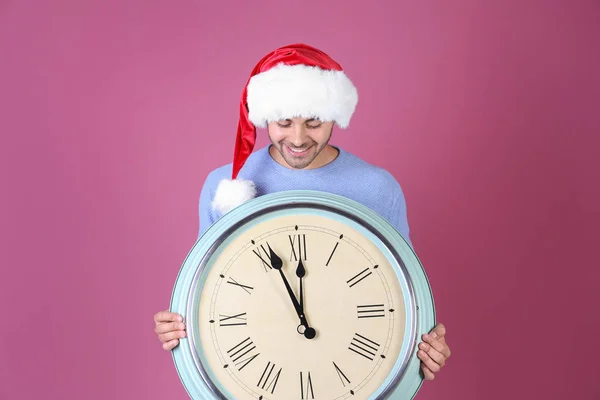 Młody człowiek w Santa hat z zegarem na kolor tła. Świąteczne odliczanie koncepcja — Zdjęcie stockowe