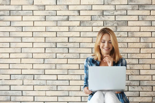 Mulher bonita com laptop moderno sentado perto da parede de tijolo — Fotografia de Stock