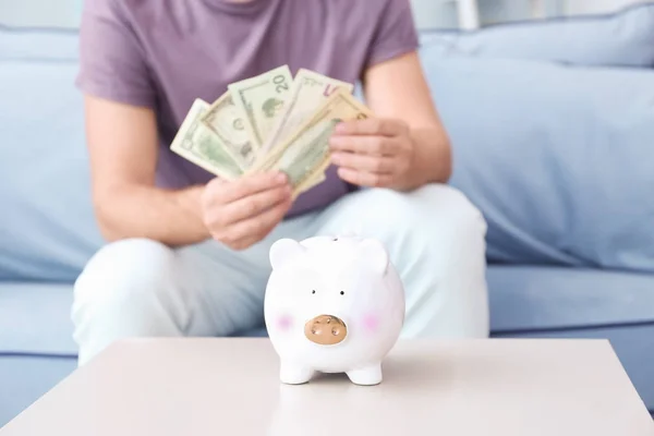 Niedliches Sparschwein auf dem Tisch und Mann mit Geldscheinen zu Hause — Stockfoto