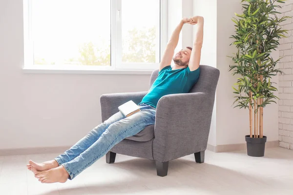 Young man with book resting in armchair near window at home — Stock Photo, Image