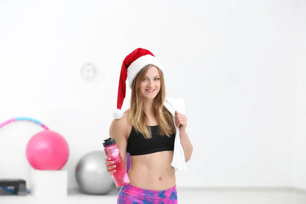 Krásná mladá žena v klobouku Santa Claus s lahví vody v tělocvičně — Stock fotografie