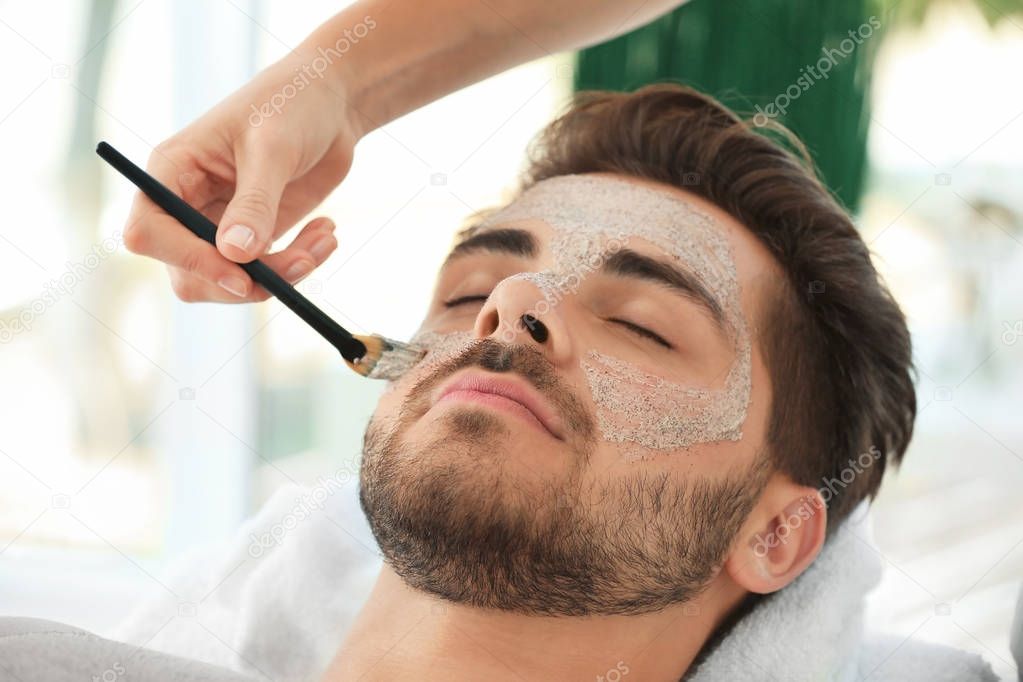 Beautician applying scrub onto young man's face in spa salon