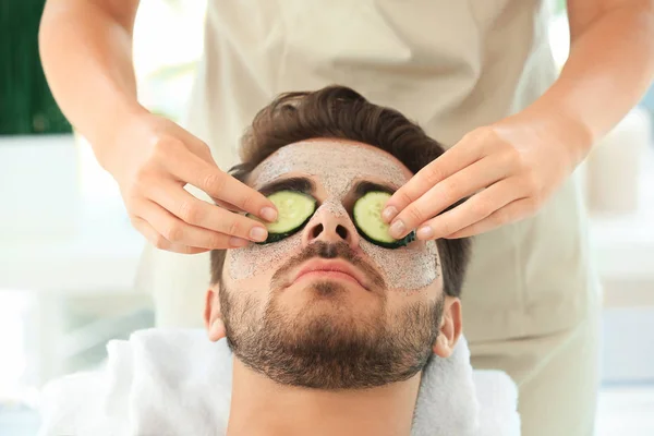 Beautician putting fresh cucumber slices on young man's face in spa salon — Stock Photo, Image