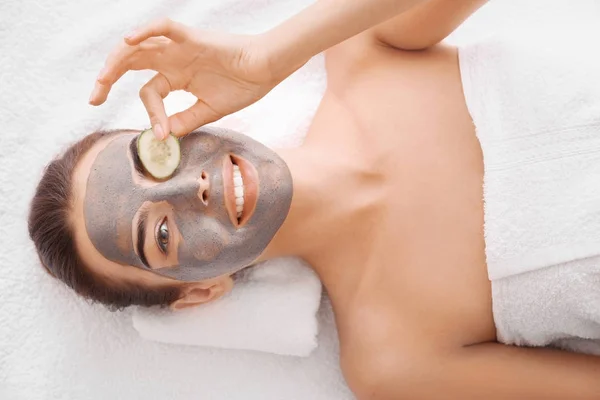 Jovem com máscara facial e fatia de pepino no salão de spa — Fotografia de Stock