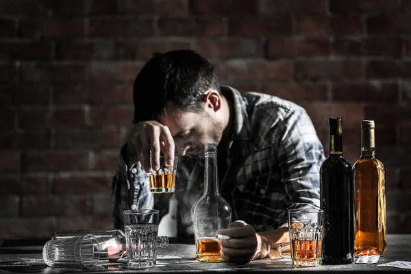 Berusad man med glas och flaskor med alkohol dryck sitter vid bord på tegel vägg bakgrund — Stockfoto