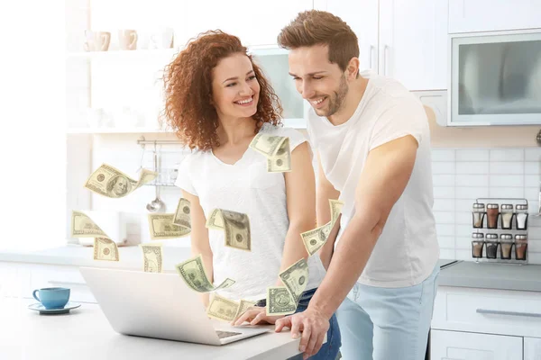 Dinero que vuela fuera de la computadora portátil mientras la pareja lo usa en la mesa en la cocina — Foto de Stock