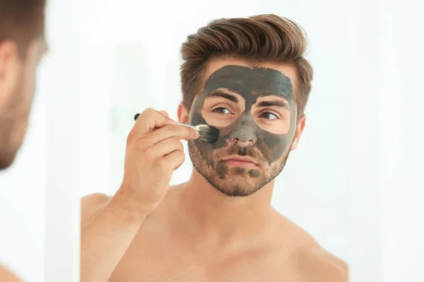 Молодой человек наносит косметическую маску на лицо в ванной комнате — стоковое фото
