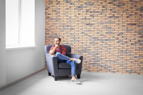 Young man sitting in armchair against brick wall — Stock Photo, Image