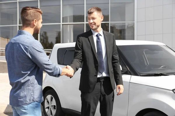 Клиент и продавец пожимают руку возле нового автомобиля на открытом воздухе — стоковое фото