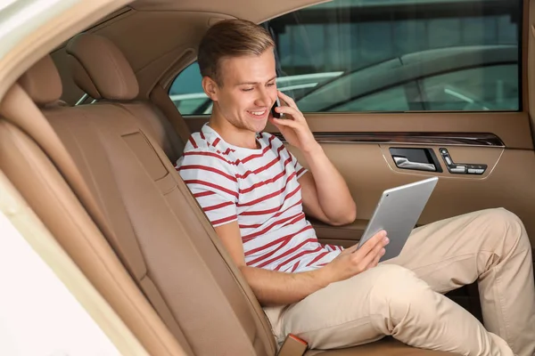 Bello giovanotto con tablet che parla sul cellulare sul sedile posteriore dell'auto — Foto Stock