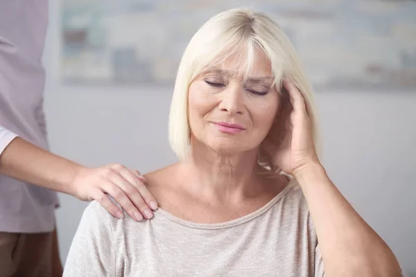 Mujer madura que sufre de dolor de cabeza, mientras que la mujer joven toma de la mano en su hombro en el interior. Concepto de cuidado de ancianos — Foto de Stock