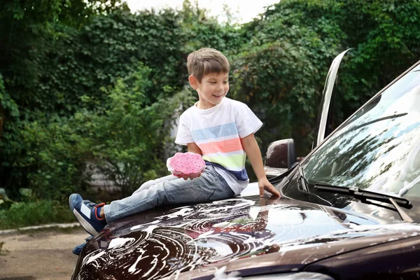 Schattige jongen wassen autootje met spons zittend op kap buitenshuis — Stockfoto