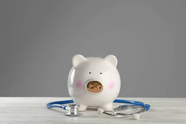 Niedliches Sparschwein mit Stethoskop auf Holztisch vor grauem Hintergrund — Stockfoto