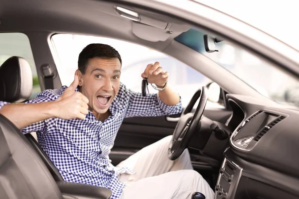 Hombre feliz mostrando la llave y el pulgar hacia arriba mientras está sentado en el coche nuevo —  Fotos de Stock