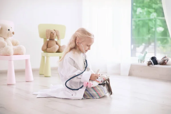 Kleines Mädchen im Arztkittel spielt mit Puppe auf dem Fußboden — Stockfoto