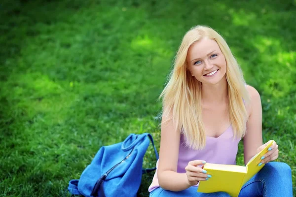 Schöne junge Frau liest Buch, während sie auf dem Rasen im Park sitzt — Stockfoto