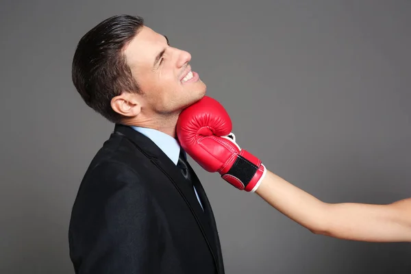 Hand of woman in boxing glove hitting businessman on grey background — Stock Photo, Image