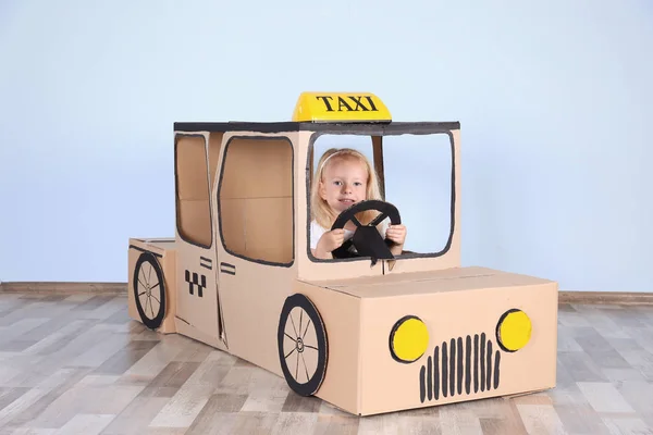 Little girl playing with cardboard taxi in light room — Stock Photo, Image