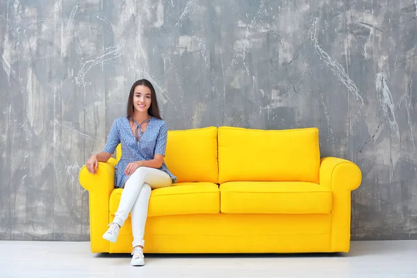 Молода жінка сидить на жовтому дивані біля сірої стіни — стокове фото