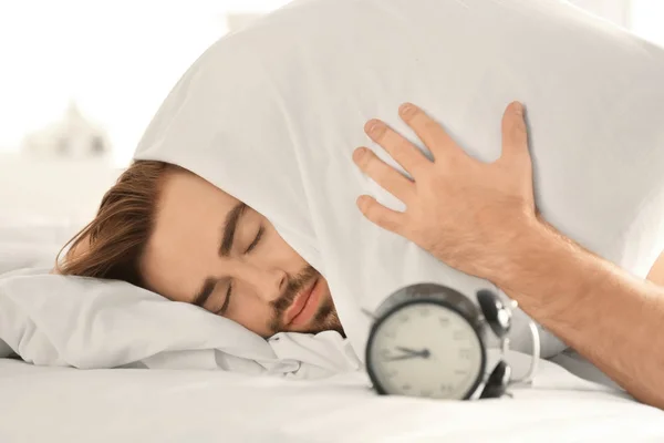 Reloj alarmante y joven que cubre la cabeza con almohada mientras trata de dormir en la cama — Foto de Stock