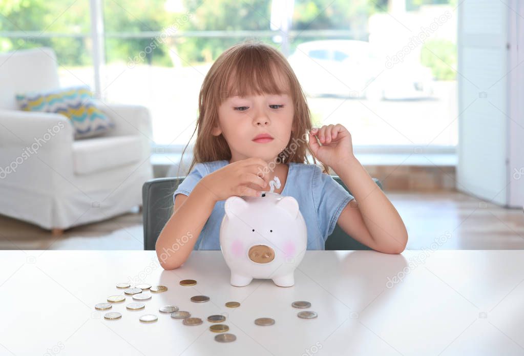 Cute little girl putting coin into piggy bank indoors