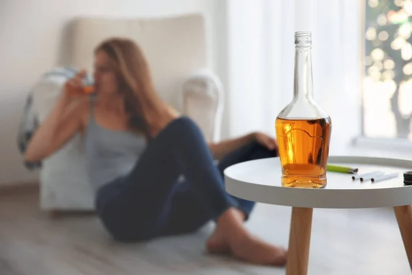 Flasche mit Alkohol auf Tisch und betrunkene Frau im Hintergrund — Stockfoto