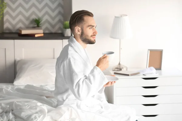 Mañana de joven guapo bebiendo café mientras está sentado en la cama en casa — Foto de Stock
