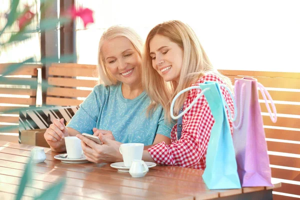 Jovem mulher usando telefone enquanto bebe café com mãe idosa no café ao ar livre — Fotografia de Stock