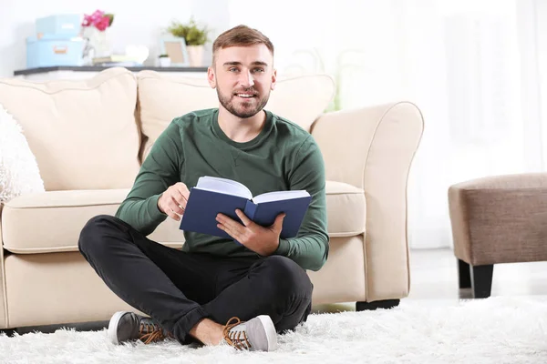 Młody człowiek czytając książkę na podłodze w pobliżu kanapie w domu — Zdjęcie stockowe