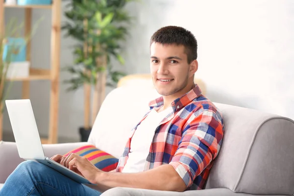 Hombre joven usando su computadora portátil mientras está sentado en el sofá acogedor en la habitación —  Fotos de Stock