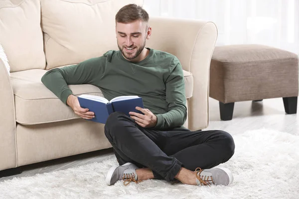 Jeune homme lisant le livre sur le sol près du canapé à la maison — Photo