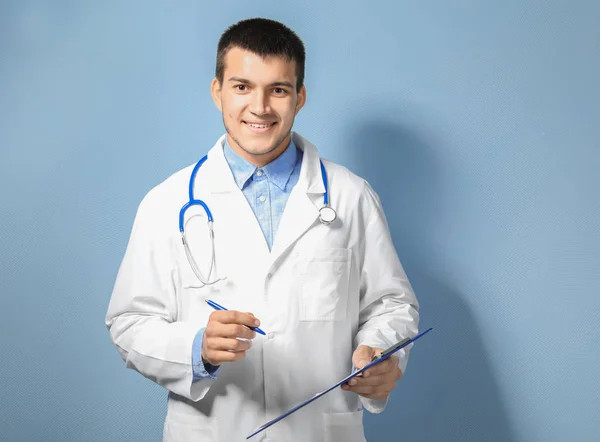 Young male doctor with clipboard against color background — Stock Photo, Image