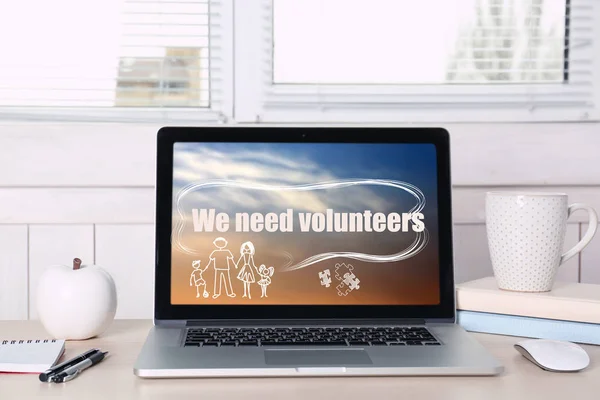 Workplace with laptop and text WE NEED VOLUNTEERS on screen in office