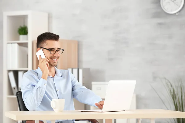 Привлекательный мужчина с ноутбуком разговаривает по телефону в офисе — стоковое фото
