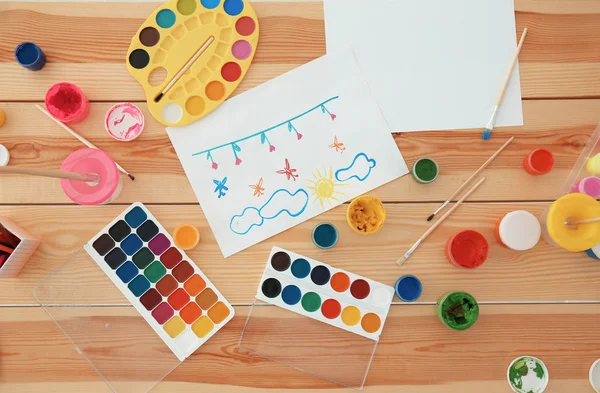 Set of paints, brushes and children's painting on wooden table — Stock Photo, Image