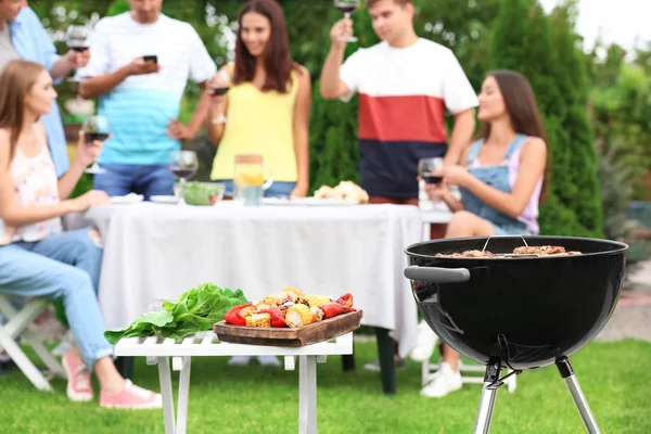 Parrilla de barbacoa, mesa con verduras preparadas y personas borrosas en el fondo —  Fotos de Stock