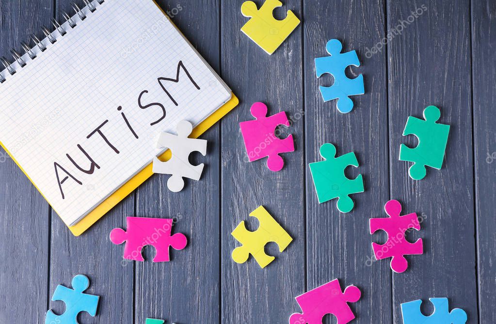 Word AUTISM written in notebook and puzzles on wooden background
