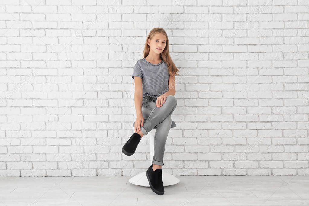 Little fashionable girl sitting on stool near brick wall