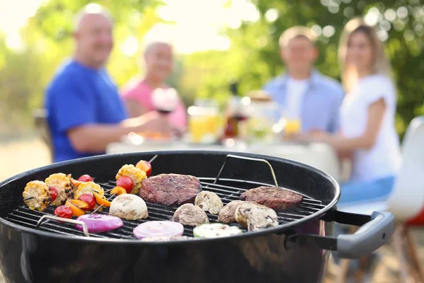 Barbecue grill with tasty steaks and happy family on background outdoors — Stock Photo, Image