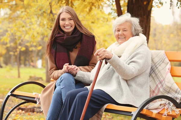 Mujer mayor con bastón y cuidador joven sentada en el banco en el parque —  Fotos de Stock