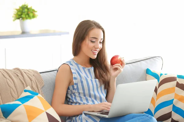 Junge Frau benutzt Laptop und isst Apfel, während sie drinnen auf dem gemütlichen Sofa sitzt — Stockfoto