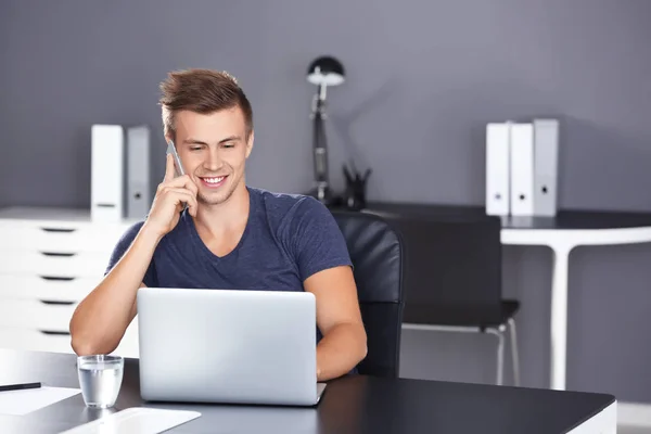 Hombre guapo con portátil hablando por teléfono en la oficina —  Fotos de Stock