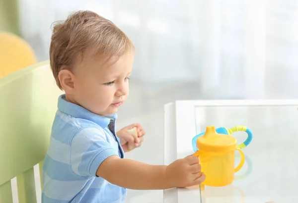 Bebê adorável sentado à mesa com bebedouro em casa — Fotografia de Stock