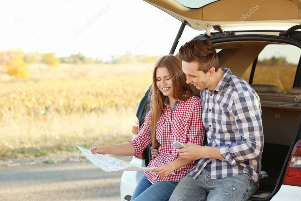 Beautiful young couple sitting with map in car trunk
