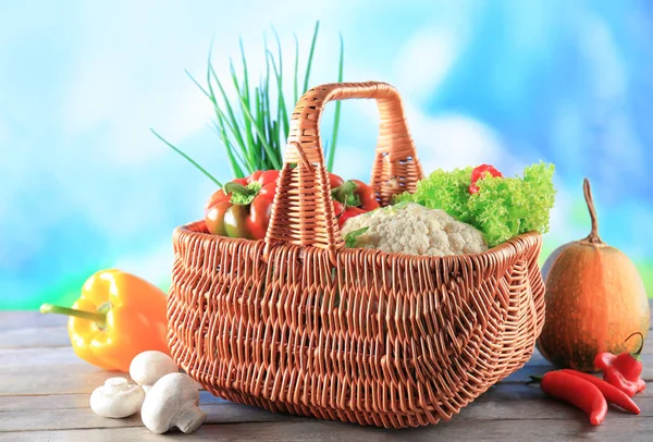 Wicker basket with different vegetables on wooden table against color background — Stock Photo, Image